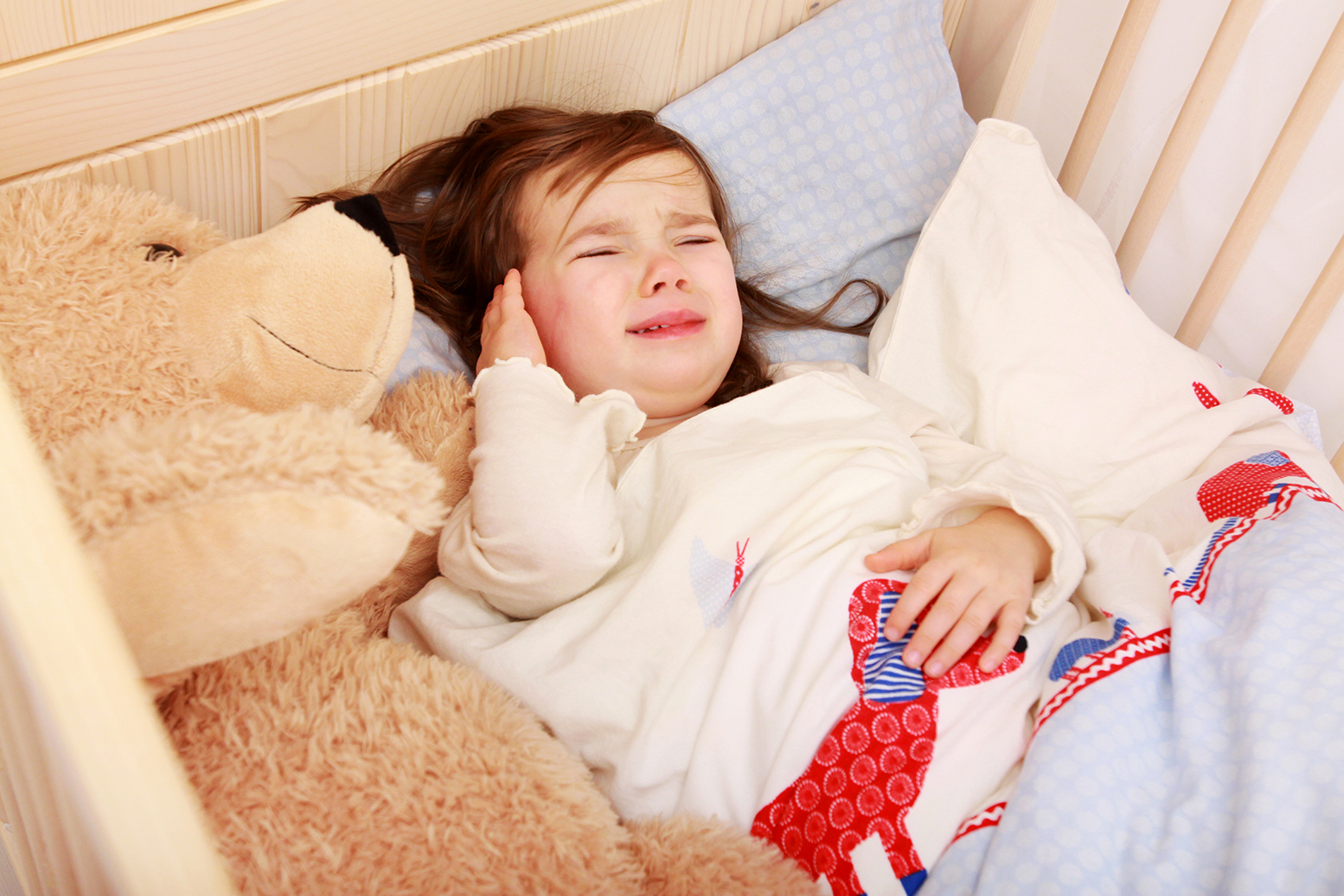 Mädchen liegt in einem Kinderbett und hält sich die Ohren, da sie Ohrenschmerzen hat.